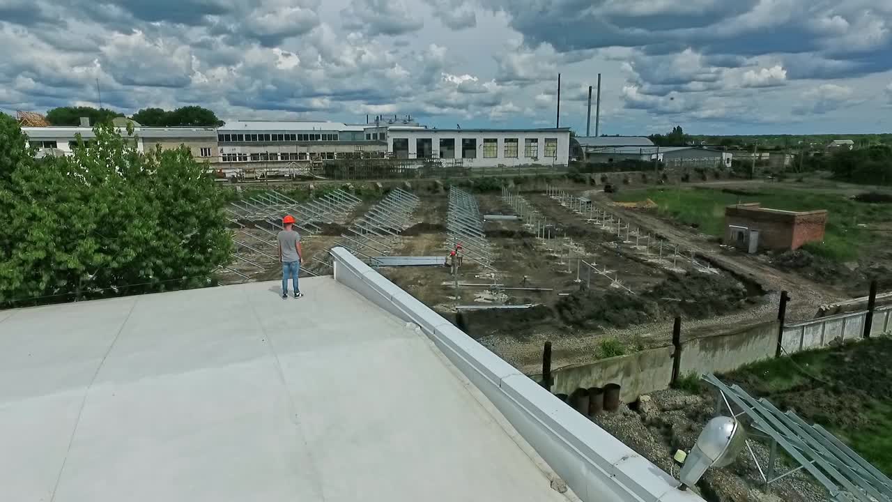 New construction of a solar farm on the field. Professional engineer standing on a rooftop of flat building and looking at the solar panels installation. Aerial view.
