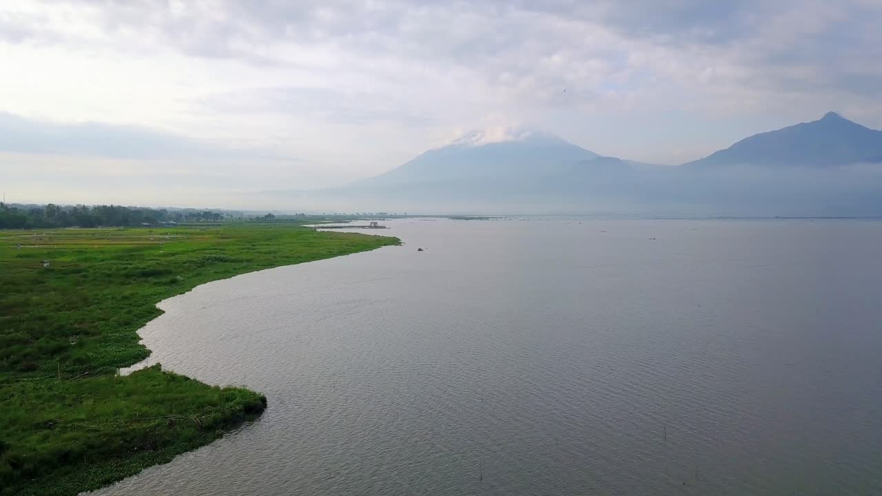 vista aérea del enorme lago y pradera verde en la orilla del lago con montaña en el fondo - lago rawa pening, indonesia