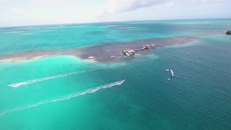 Dos-Hombres-Kitesurfistas-Cruzan-El-Agua-Del-Mar-Volando,-Disparos-De-Drones-Palafito-Los-Roques