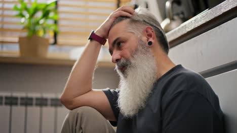 close up a serious elderly man with gray hair and a lush beard looks with downcast eyes and thinks about his life sitting on the floor in the kitchen and takes his head