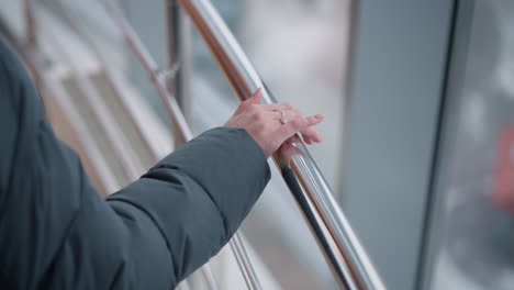 close-up of woman hand adorned with ring resting on rail while walking, with glass building reflecting surroundings in background, capturing elegance, modern style, and contemplative atmosphere