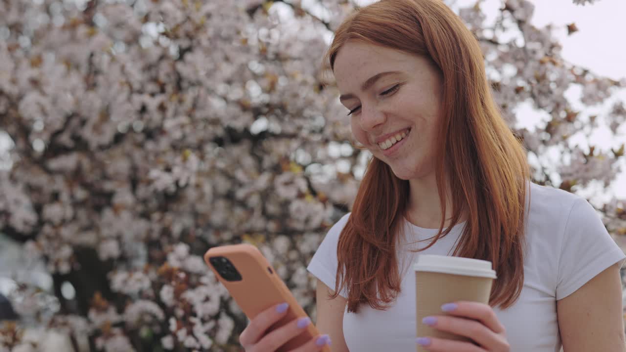 Woman using phone in a park with cherry blossoms and coffee