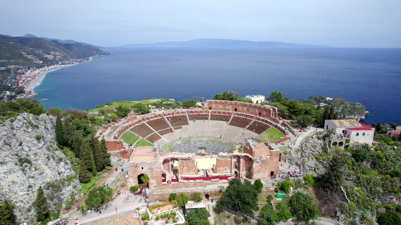 Um drone circula o Teatro Antico di Taormina, revelando seus assentos curvos, paredes de palco em ruínas e visitantes com o azul do Mar Jônico ao fundo