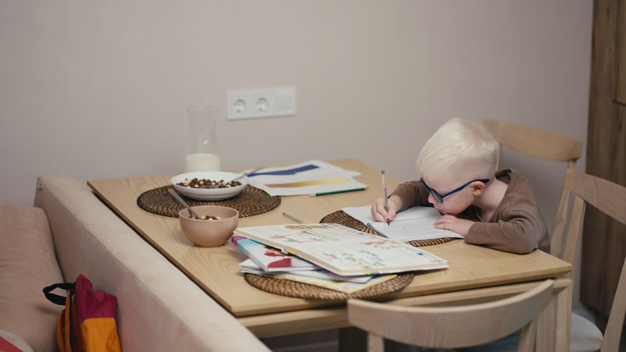 Little albino boy with white hair color in blue glasses doing homework and writing his notebooks while preparing for the next day at school in a modern kitchen in an apartment during the day