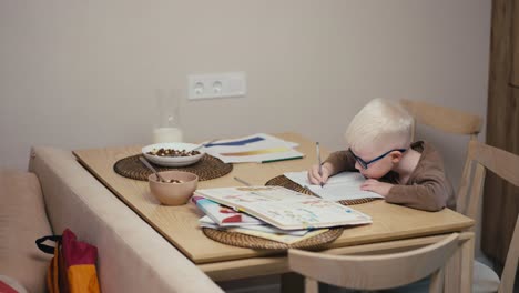 Little-albino-boy-with-white-hair-color-in-blue-glasses-doing-homework-and-writing-his-notebooks-while-preparing-for-the-next-day-at-school-in-a-modern-kitchen-in-an-apartment-during-the-day