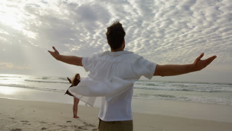 vista trasera de una pareja joven de pie con los brazos extendidos y mirando al mar en la playa 4k