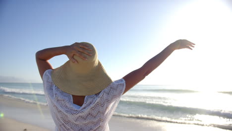 Una-Joven-Birracial-Disfruta-De-La-Playa,-Con-Los-Brazos-Extendidos-Y-Un-Sombrero-De-Ala-Ancha-En-La-Cabeza