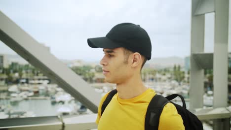 young attractive trendy young man wearing a cap and a backpack walking on a bridge on a sunny day with an urban city background