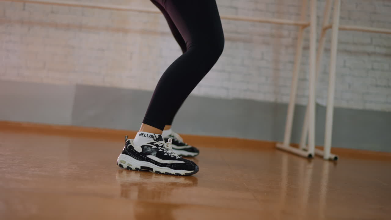 Side view of young girl in black leggings and canvas sneakers squatting down with hand on waist in studio showing expressive body posture fitness rhythm athletic training movement confident energy