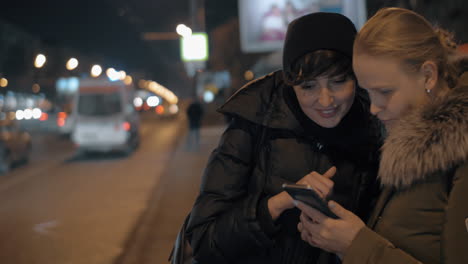 Mujeres-Viendo-Algo-En-El-Móvil-Mientras-Esperan-En-La-Parada-De-Autobús-Por-La-Noche