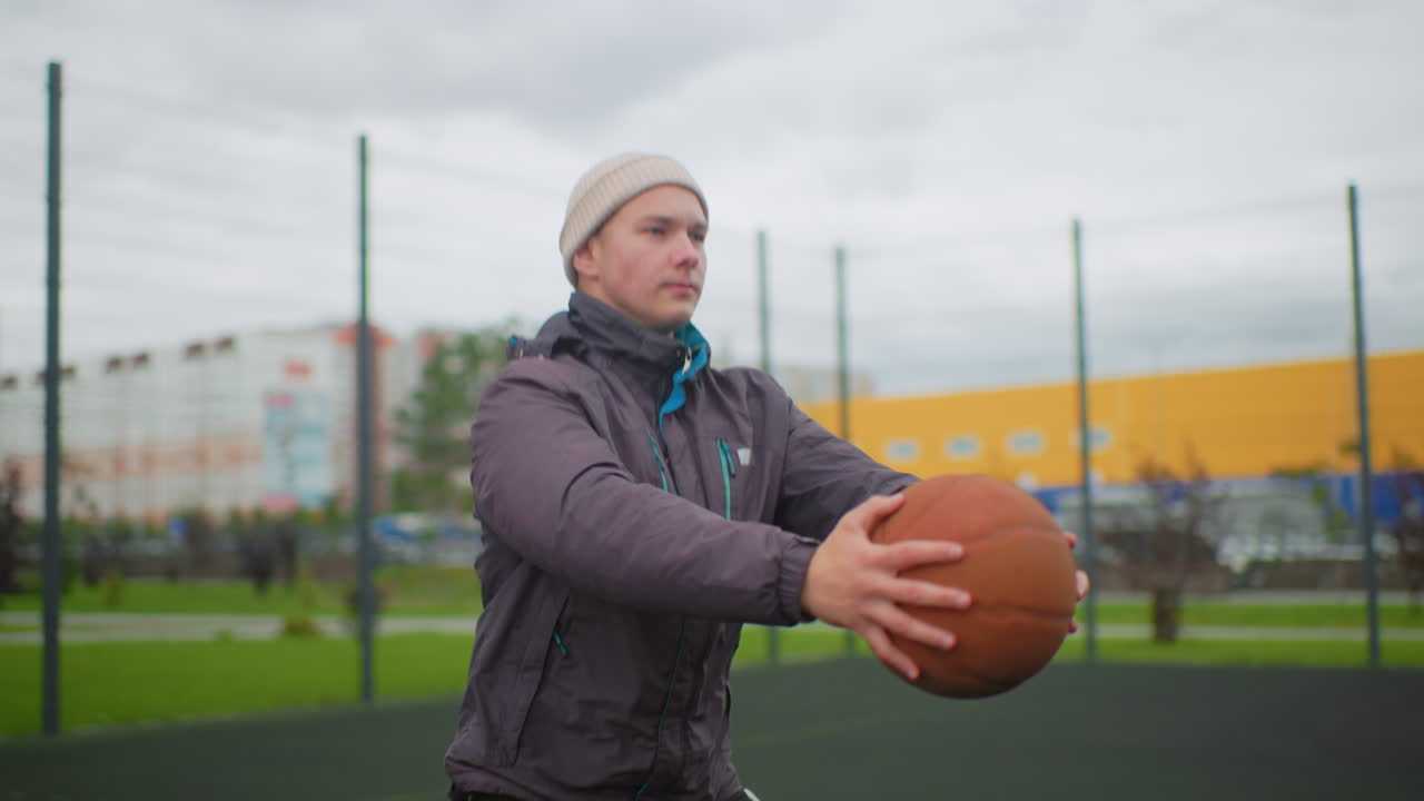 Hvid mand holder basketball udendørs på bymæssig indhegnet bane, iført vinterjakke og hue, overskyet himmel og farverig lagerbygningsbaggrund, står stille med hænderne på bolden, eftertænksom stemning