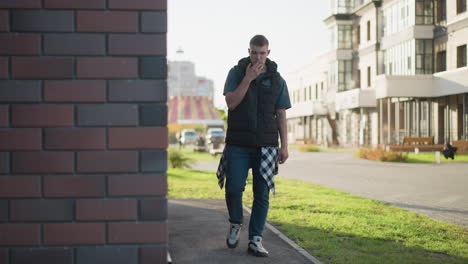 young man walks alone along paved sidewalk smoking cigarette near urban building during daylight with blurred modern structures in background and green grass framing edge of cityscape