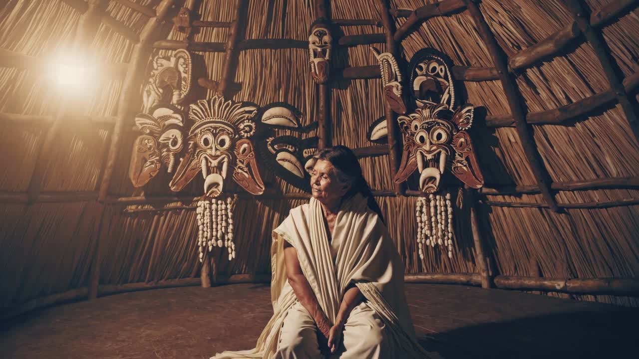 Elderly woman in traditional attire sits contemplatively in a dimly lit hut adorned with vibrant masks, showcasing cultural heritage and deep-rooted traditions in a serene atmosphere