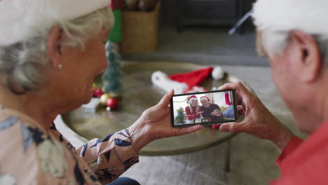 pareja caucásica mayor sonriente usando un teléfono inteligente para una videollamada de navidad con la pareja en la pantalla