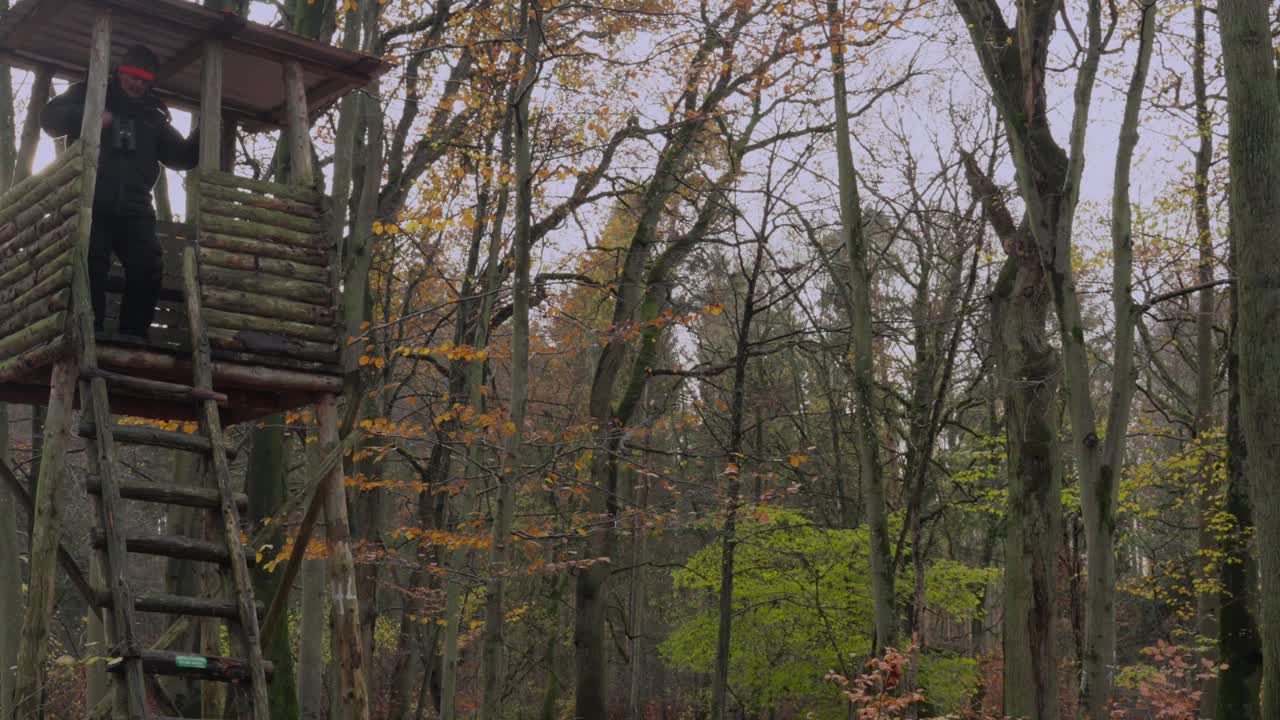 un cazador en un grueso traje de invierno se sienta en un soporte alto, luego se levanta y baja la escalera y corre fuera de la imagen, lleva un rifle de caza con un silenciador sobre el hombro