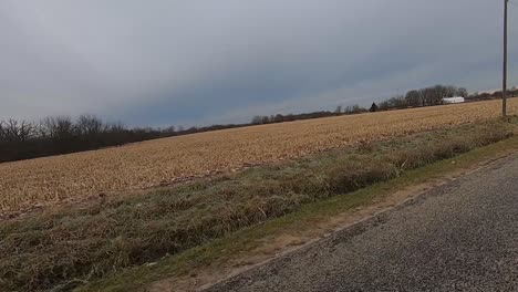 POV-through-drivers-side-window-while-driving-down-a-rural-county-road-in-late-afternoon-past-harvested-fields