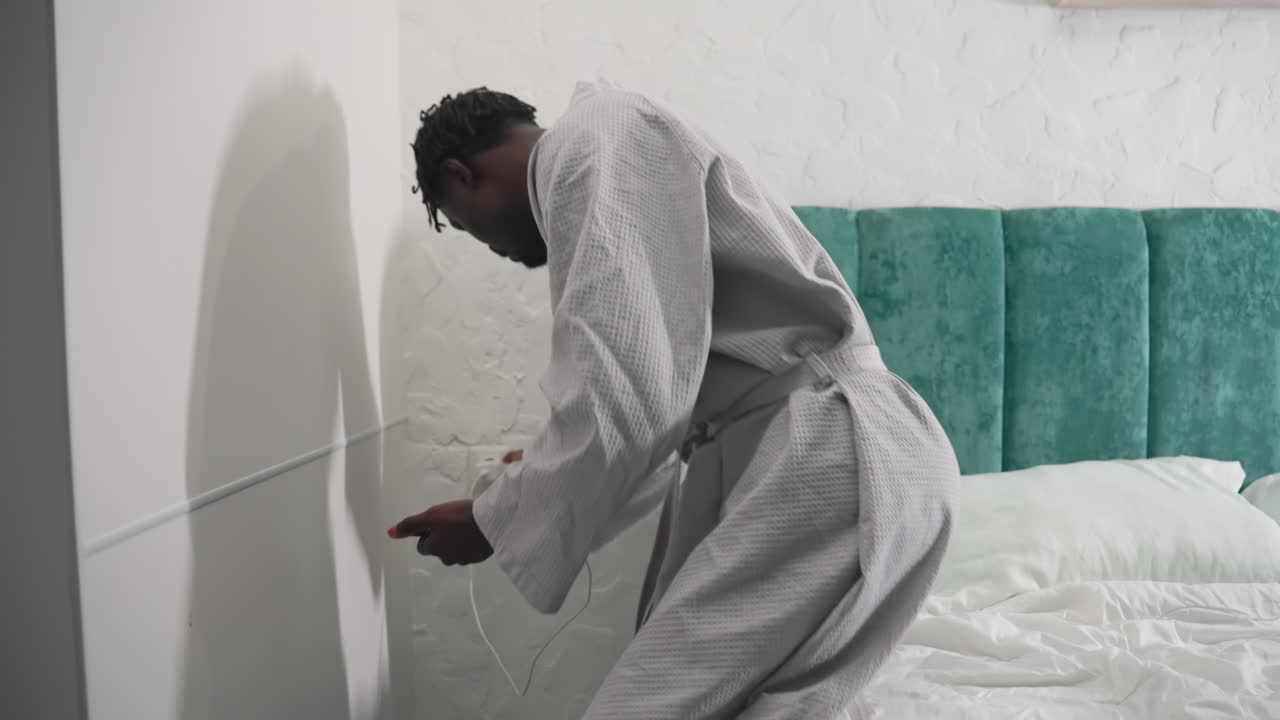Youth in bathrobe standing by bed in cozy bedroom adjusting bedside light. Calm morning scene in a modern home with green headboard. Relaxed atmosphere as they prepare for the day ahead