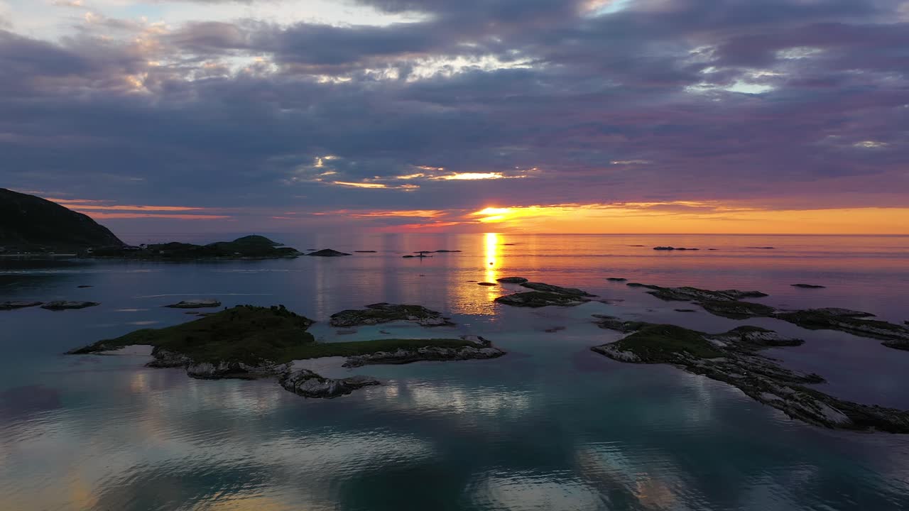 Aerial view low over small islands and mirroring water, towards the colorful, midnight sun, dark clouds, during sunset, at the Arctic ocean, in Sommaroy, Norway - low, dolly, drone shot