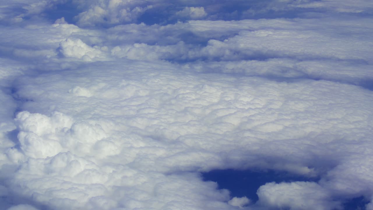 A high-altitude aerial perspective looking down at a vast, textured layer of white cumulus clouds under a clear blue sky, captured from a passenger plane during flight