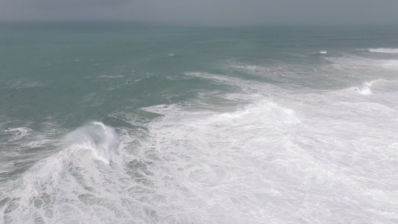 Aerial drone shot of big waves coming into shore on a day with giant waves in Nazaré, Portugal, Europe. Jet ski riders and surfers in Atlantic Ocean. Stormy rough surf conditions