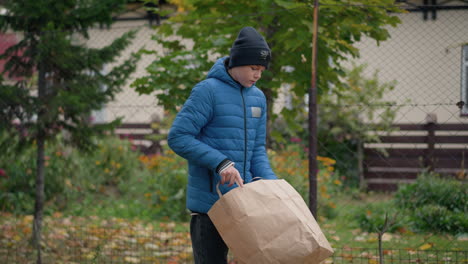 little boy walking along dry leaves thoughtfully, turns paper bag upside down, joyfully pouring dry leaves on the ground with a smile, surrounded by autumn colors and vibrant greenery