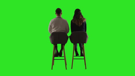 rear view studio shot of man and woman sitting on stools facing away from camera against green screen