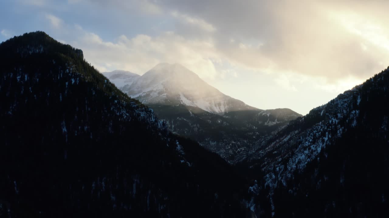 Static aerial drone shot of a winter landscape of Mount Timpanogos in the background surrounded by a pine tree forest during sunset from the frozen Tibble Fork Reservoir in American Fork Canyon, Utah