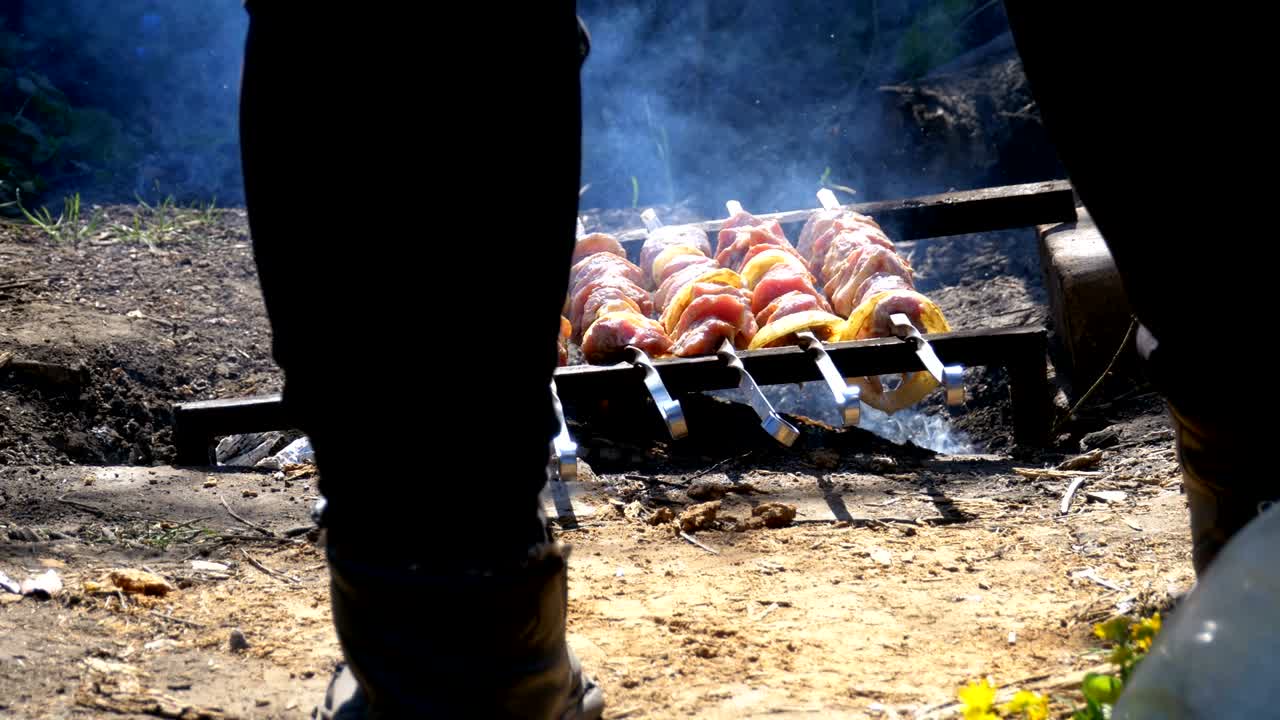 A view of the legs of a man cooking Shish kebab on skewers over a fire in the countryside in the village