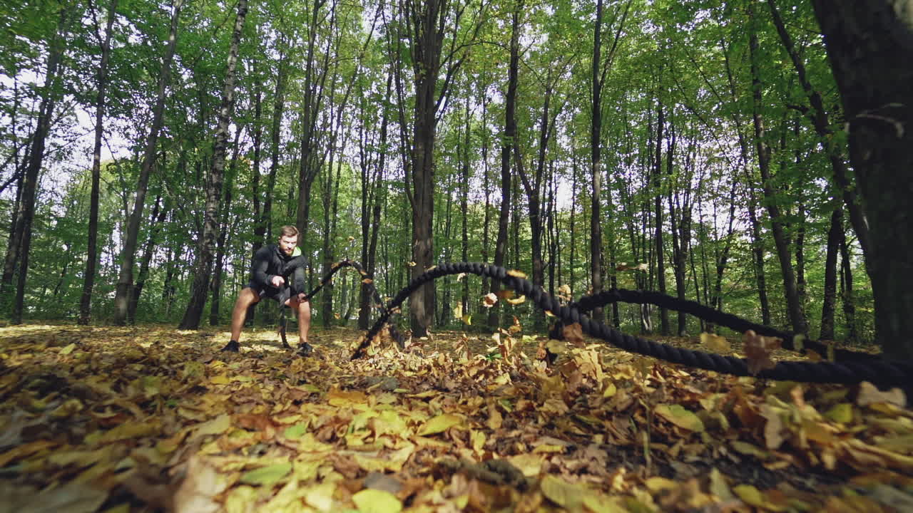 Training with battle ropes in the forest in autumn. Sportsman doing waves with ropes outdoors. Front view. Slow motion. Healthy lifestyle.