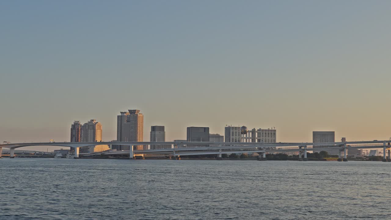 Wide-angle shot of the Rainbow Bridge and the city skyline with the sun setting, casting a golden light on the water