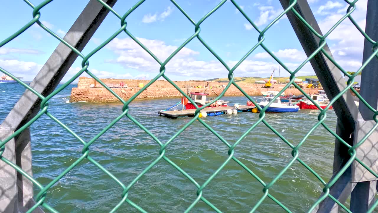 Fishing boats move across a harbor, viewed through a green chain link fence on a pier. Bright daylight, steady camera, coastal town background
