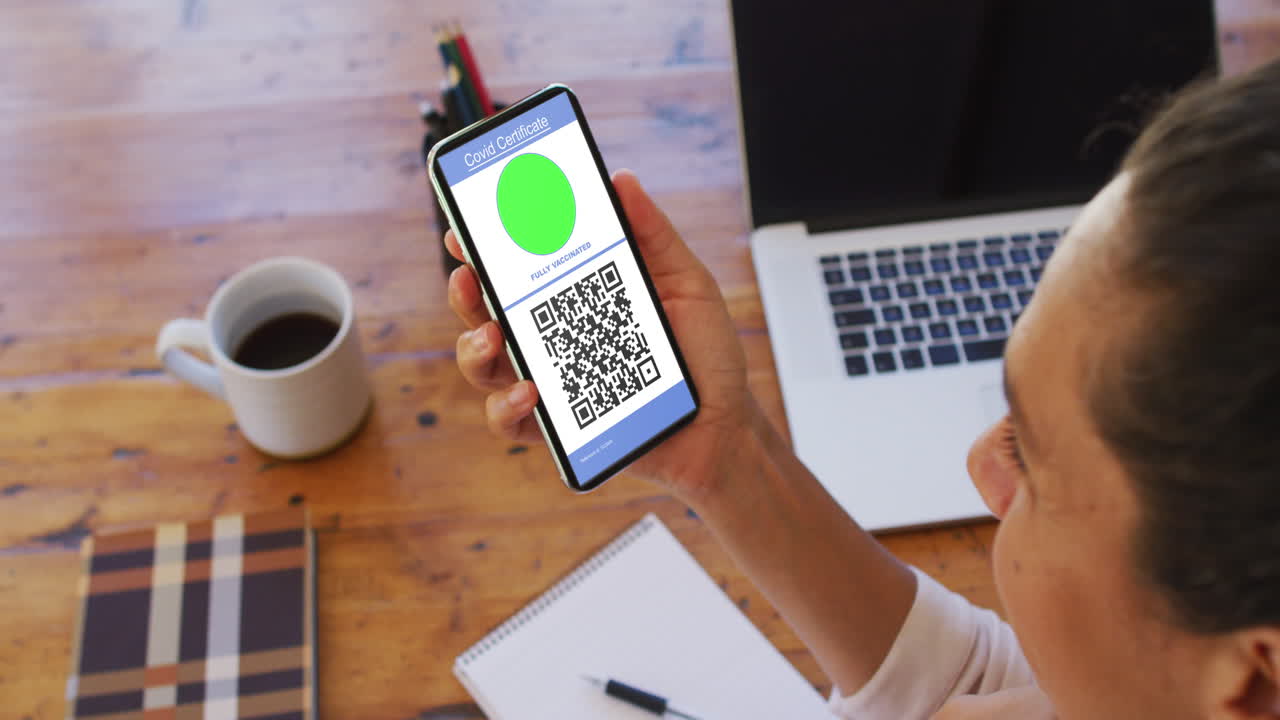 Woman at desk holding smartphone with covid vaccination certificate and qr code on screen