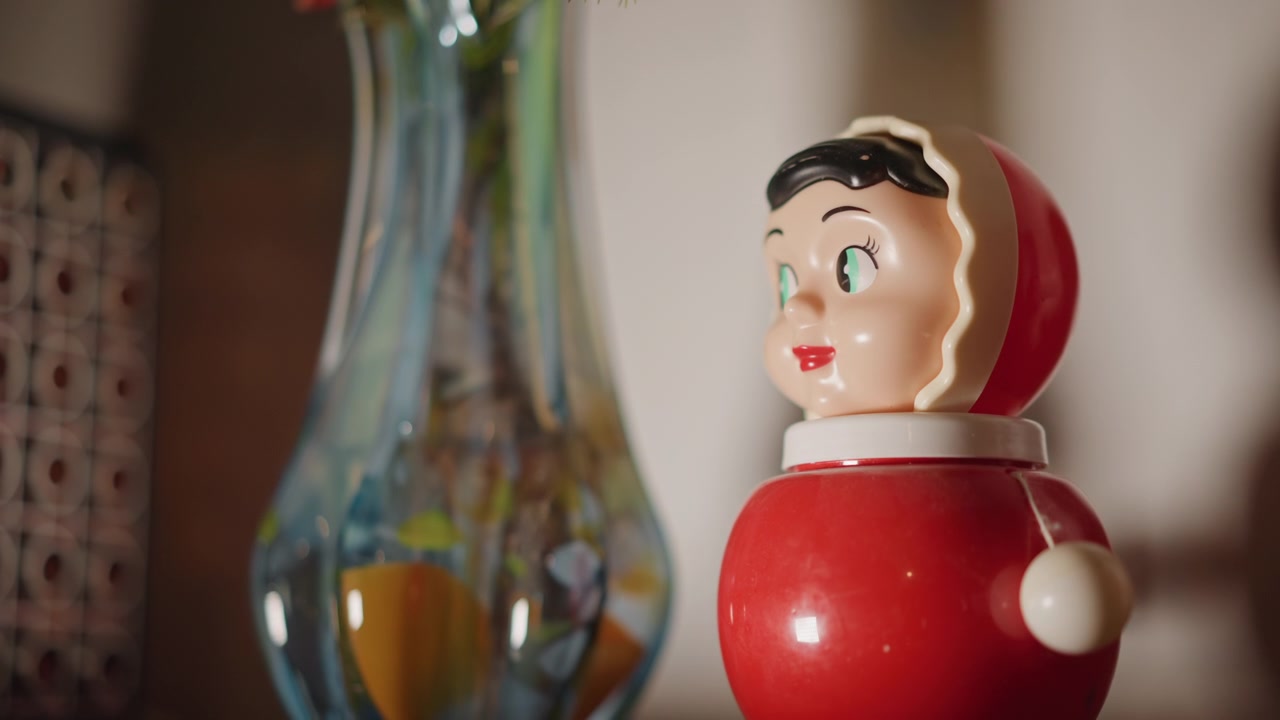 Vintage red toy beside glass vase, closeup shot of retro bobble doll on wooden shelf, warm window light, shallow depth of field, soft bokeh, glossy paint details, subtle wear and patina, cozy interior