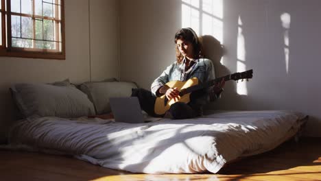 Relaxed-mixed-race-woman-sitting-on-bed-playing-acoustic-guitar-in-sunny-cottage-bedroom