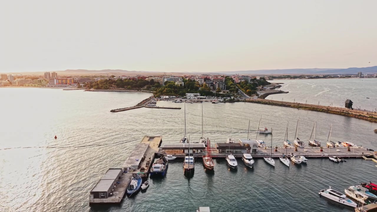 Vista de uma marina com barcos durante o pôr do sol perto de uma cidade costeira com edifícios e árvores do outro lado da água