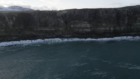 Vista-Del-Acantilado-De-Falda-Escocesa-En-Escocia-Con-Montañas-Cubiertas-De-Nieve-En-La-Distancia-Y-Olas-Rompiendo-En-Tierra