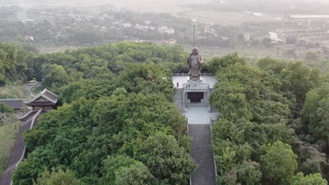 Drone-aerial-view-in-Vietnam-flying-away-from-a-buddha-statue-on-a-green-tree-covered-valley-in-Ninh-Binh-at-sunset