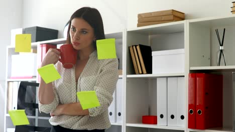 businesswoman with cup in hands considering stickers on glass in office