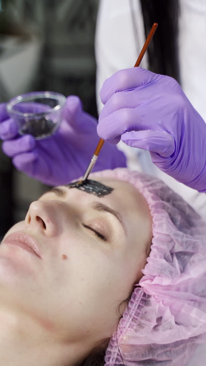 A beautician applies a black mask on the face of the client's face in a beauty salon. Skin care. Beauty industry Vertical video