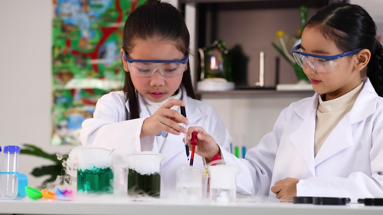 Dua gadis Asia mengenakan jas lab menggunakan pipet untuk eksperimen kimia berwarna-warni dengan gelas kimia.