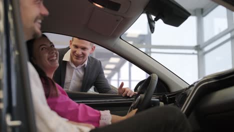 young car salesman showing to young couple new automobile at dealership salon.