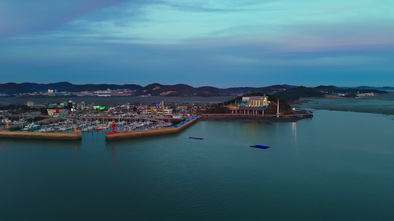 Ripresa aerea con drone in parallasse della Funivia Marina di Jebudo in movimento dalla stazione di Jeongok all'ora blu, con marina, faro rosso e calme acque costiere coreane.