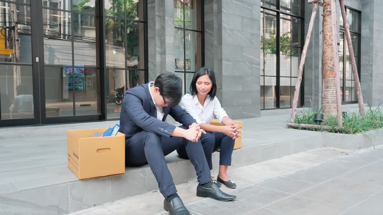 dos hombres de negocios que son despedidos se sientan frente al edificio de oficinas con una caja de pertenencias personales. recesión empresarial, renunciar a un trabajo, reducción de personal y concepto de desempleo. cámara lenta
