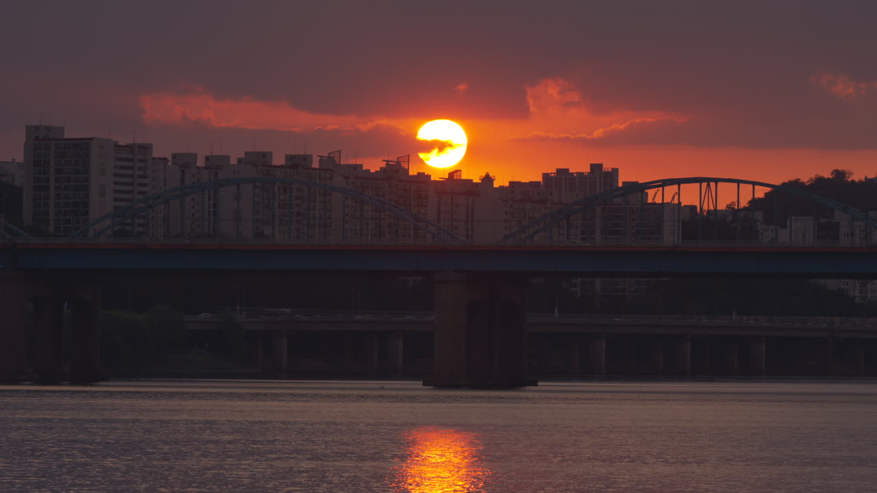Sunset over Seoul’s Dongjak Bridge with the Han River glowing below. High-rise apartments silhouette against the fiery sky as the sun dips, blending urban landscape with natural beauty