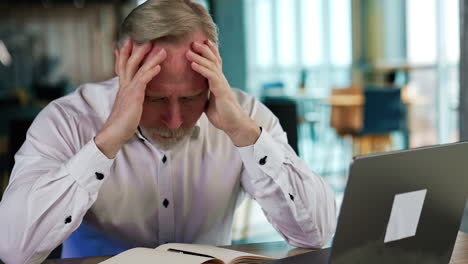 troubled senior man sits at desk holding by the head. tired overworking businessman in office. blurred backdrop.