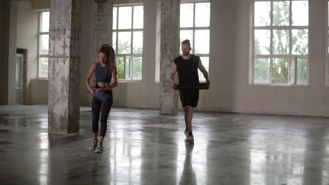 preparing for training in empty studio with panoramic windows. young woman with dreadlocks and man rolls apart yoga mat in studio. freedom, health lifestyle concept