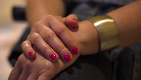 manos de una mujer con uñas pintadas de rosa con una amplia pulsera de oro descansando en su regazo