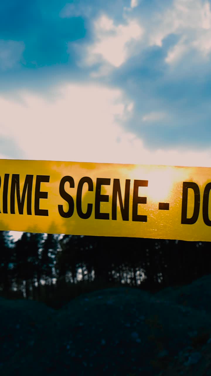 Close up view panning or moving along a yellow colored police investigation Crime Scene forensics tape ribbon that is backlit by sun setting in a quarry mine area with blue sky, white clouds and trees