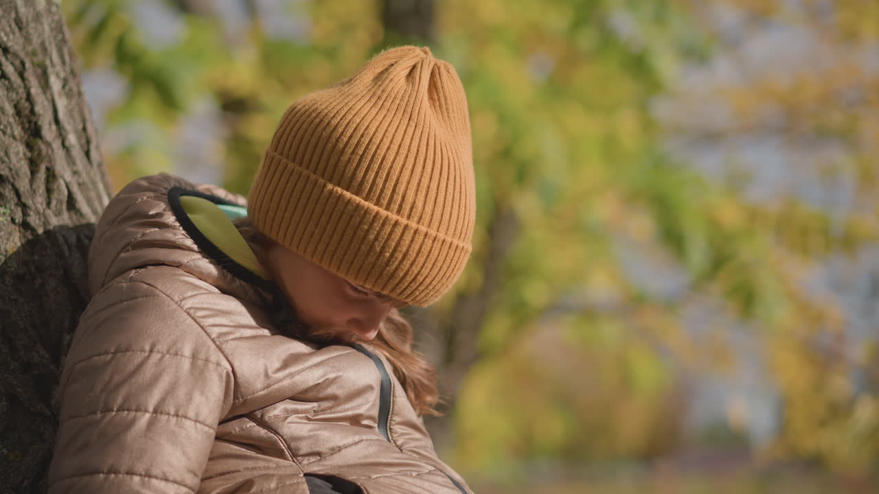 young girl in mustard knit hat and puffy coat leans against tree trunk, resting calmly in golden autumn park with soft sunlight casting gentle glow on leaves and serene expression on her face