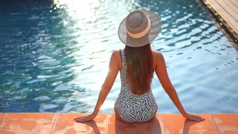 a woman sitting on the edge of a resort pool with her back to the camera looks across the surface of the swimming pool